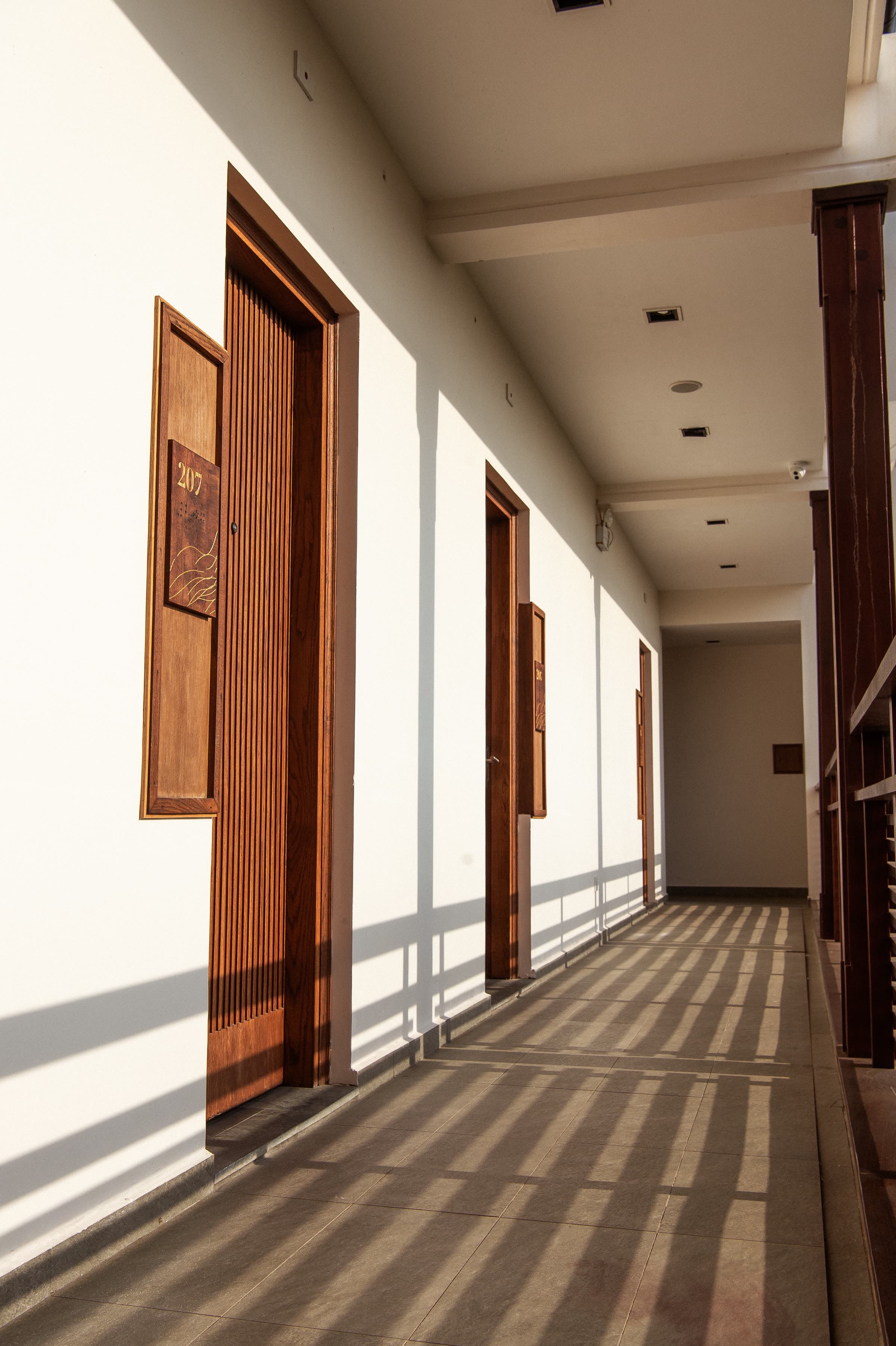 Sunlight falling across the guest-room corridor at Nghê Prana, timber doors along a white wall — a Hoi An riverside hotel