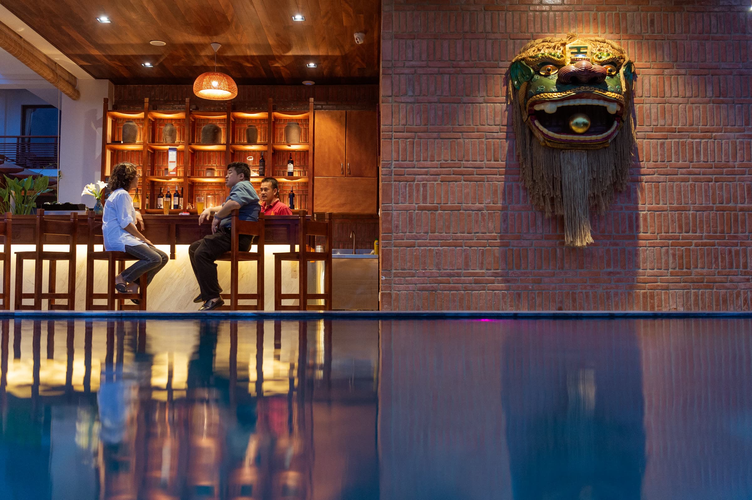 Guests with drinks at the poolside bar at dusk, the gilded Nghê mask on the brick wall behind — The Corn Restaurant & Bar at Nghê Prana, Hội An