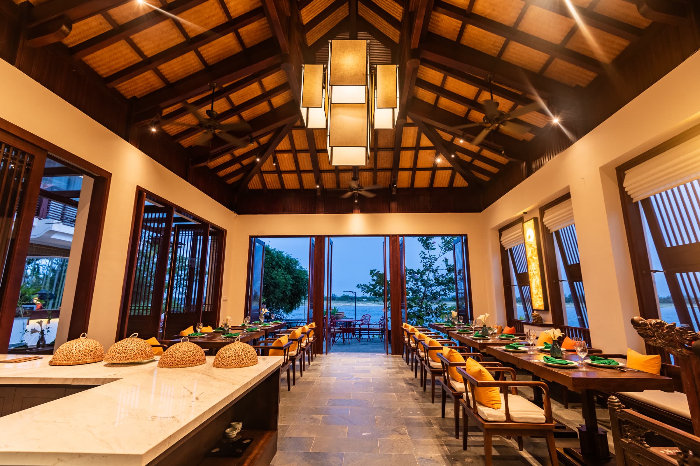The Corn Restaurant at Nghê Prana — long timber tables under a vaulted roof and a paper lantern, the doors at the far end open to the Thu Bồn River
