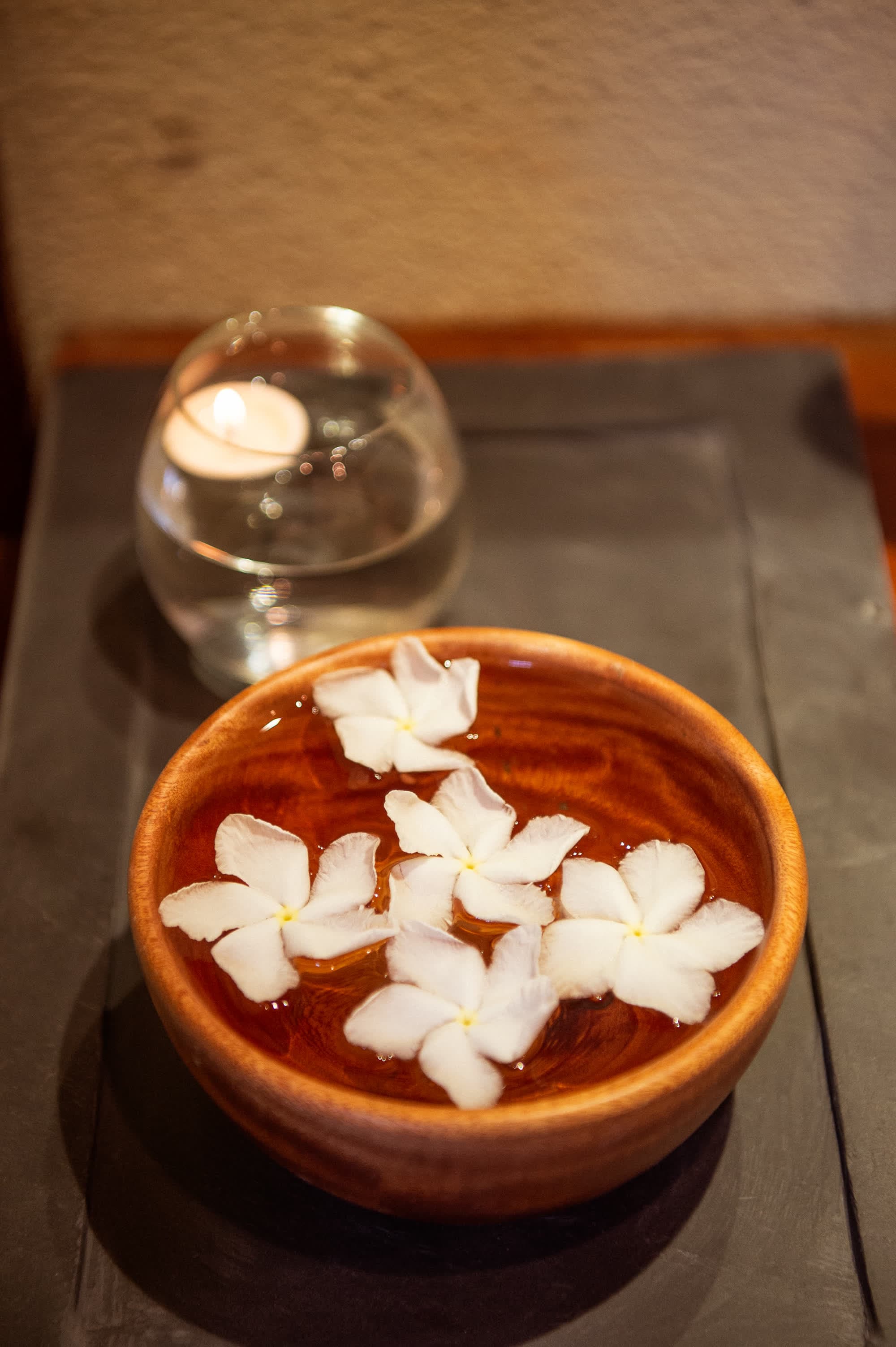Frangipani flowers floating in a wooden bowl beside a lit candle at Nghê Prana spa