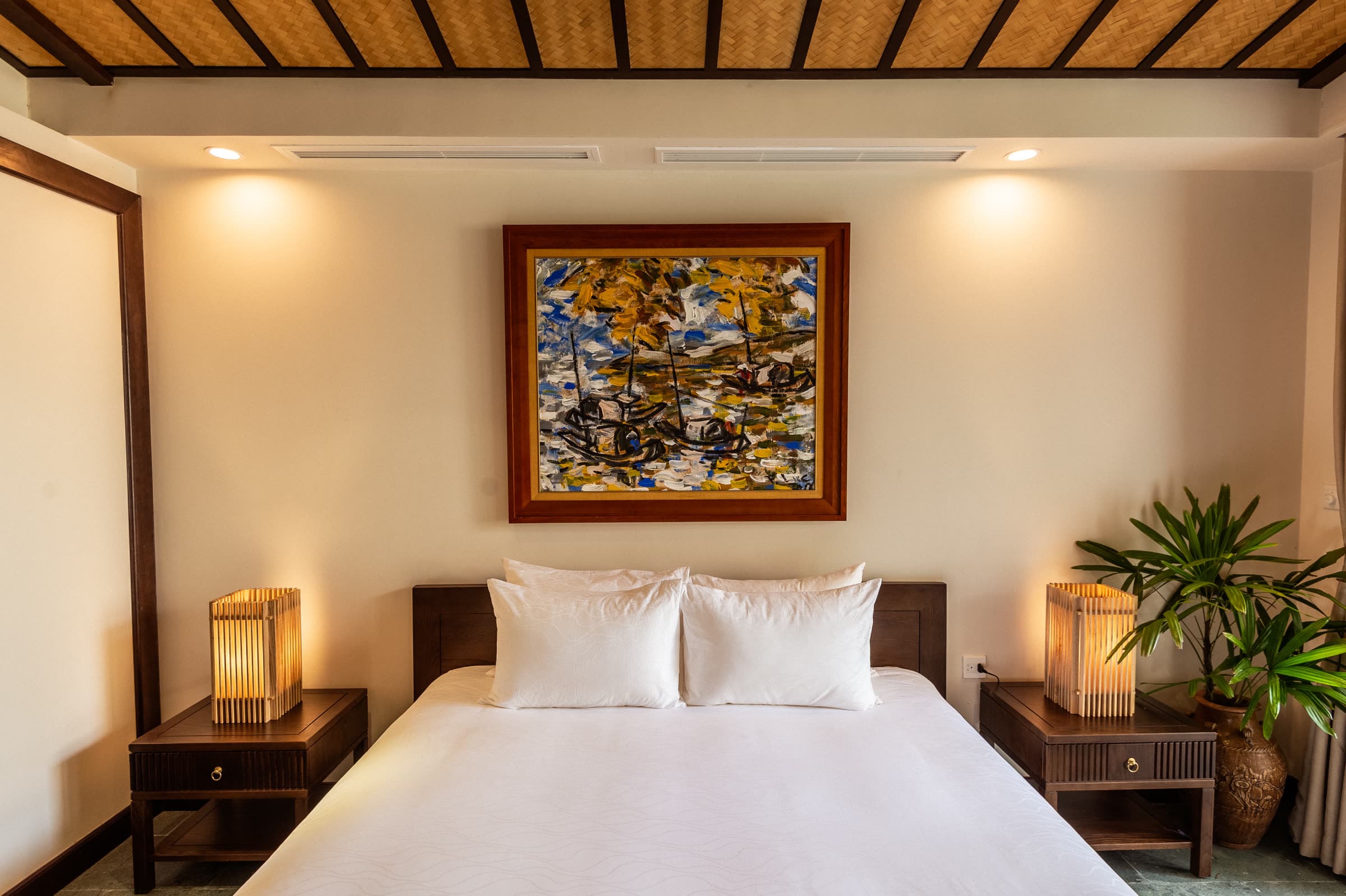 Junior Suite bed flanked by woven bedside lanterns beneath a framed Vietnamese lacquer painting