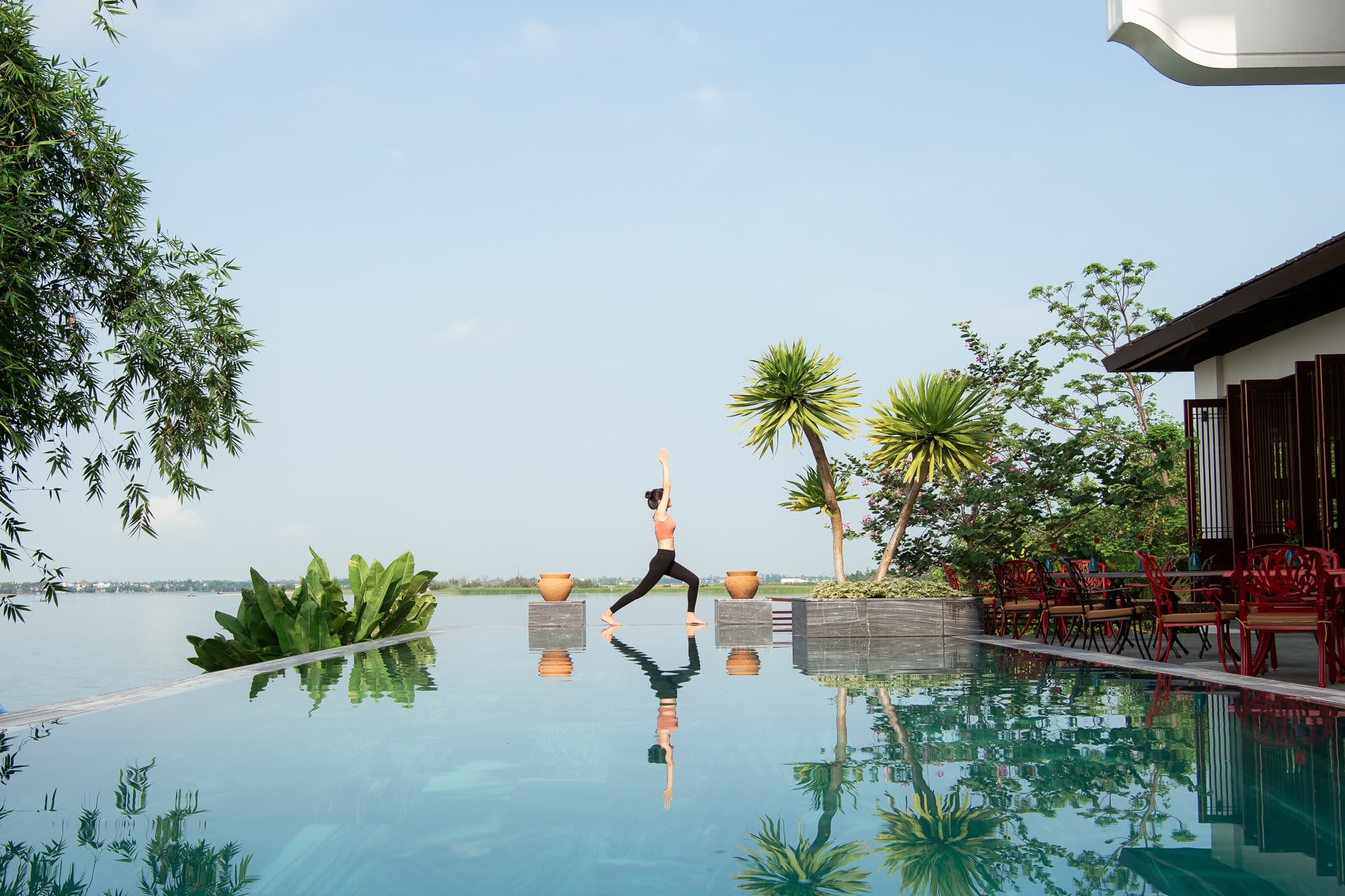 Warrior pose on the pool deck at Nghê Prana, reflected in the infinity pool with the Thu Bồn River beyond