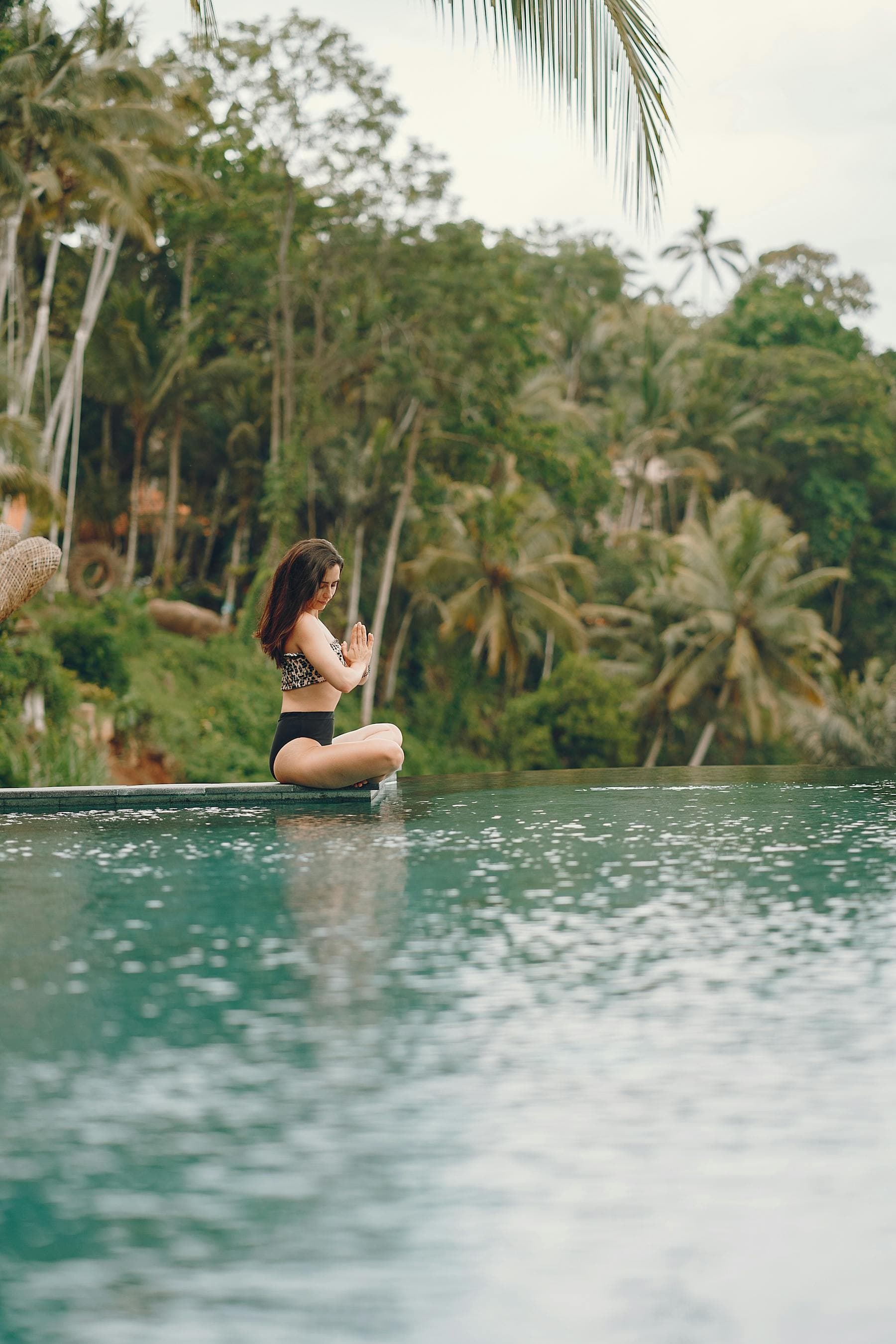 Woman meditating in lotus pose beside a tropical infinity pool surrounded by palms, illustrating the 2026 Asian wellness tourism boom.