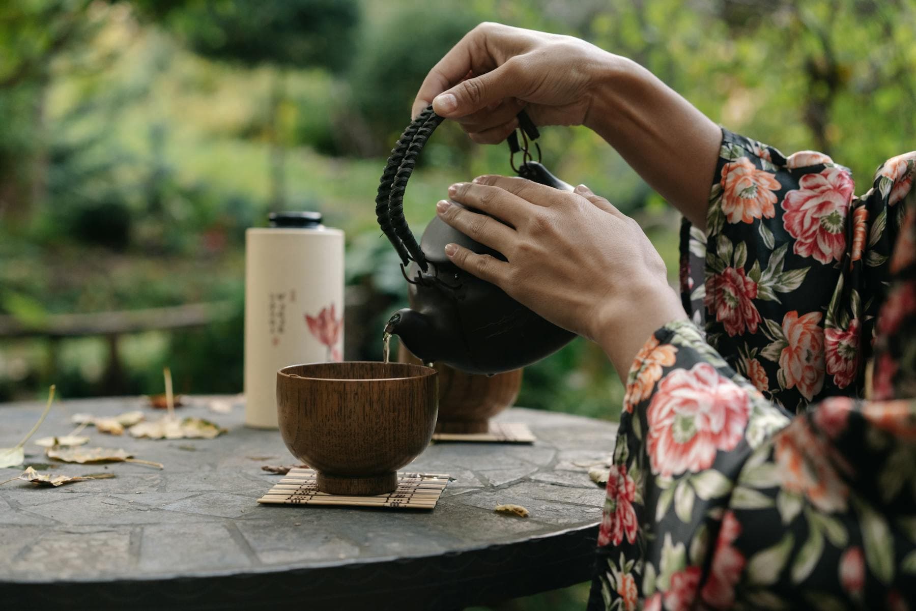 Asian woman in floral robe pouring hot tea from a cast iron kettle into a wooden bowl outdoors, a Vietnamese morning tea ritual that predates the Chinamaxxing wellness trend by centuries