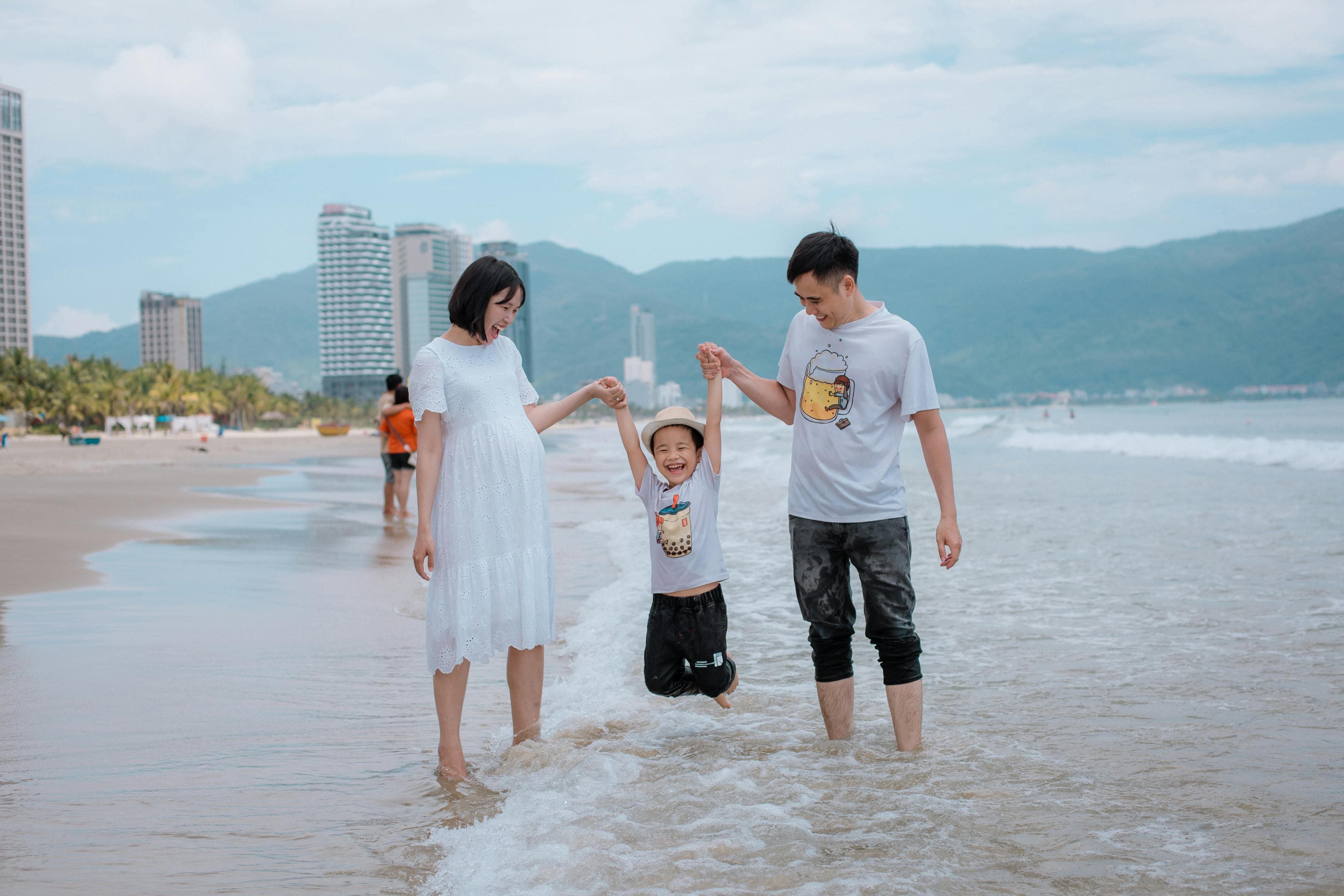 Young Asian family with child jumping happily in shallow waves on Da Nang beach — central-Vietnam family travel beach scene near Hội An