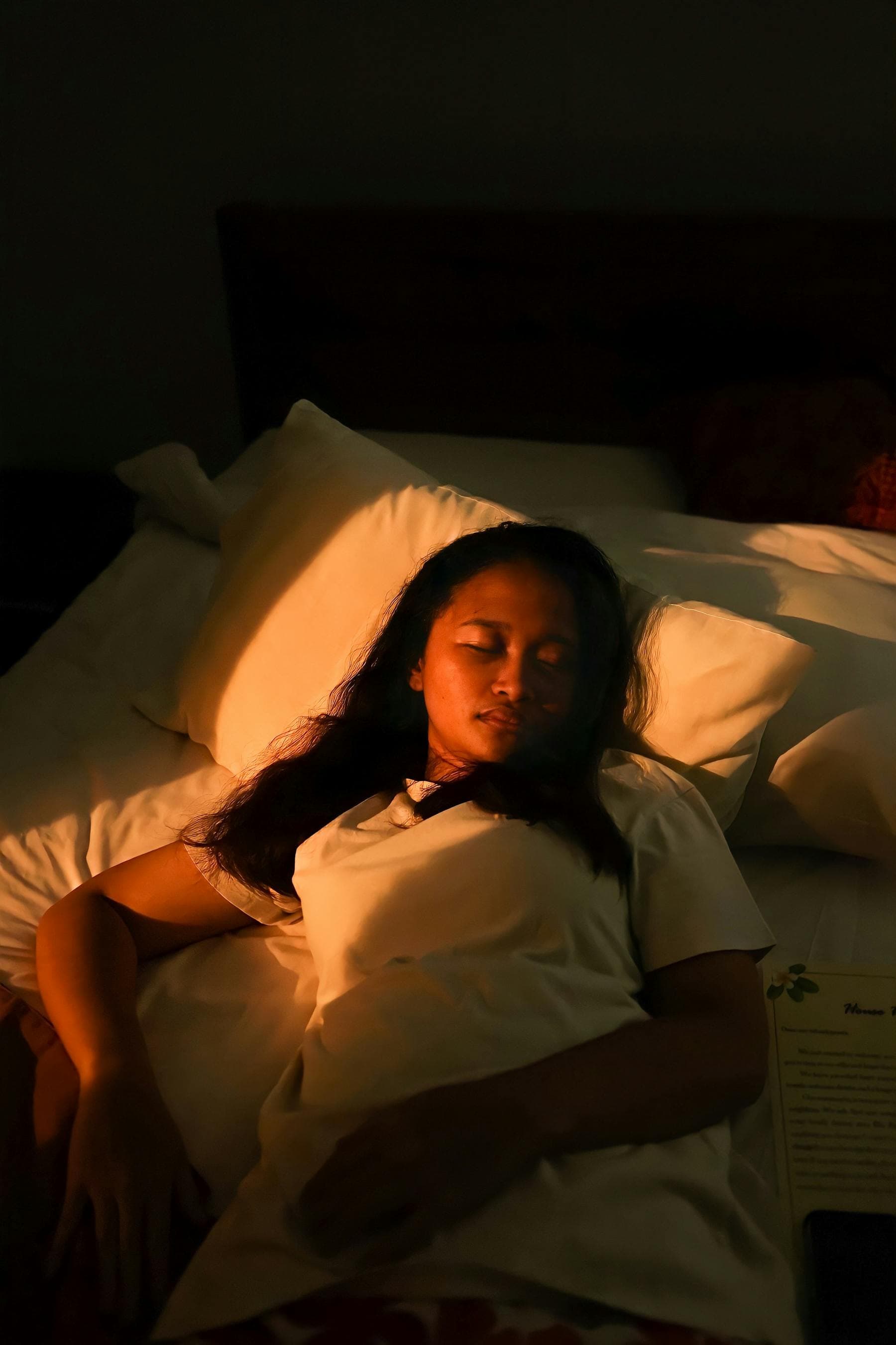 Asian woman sleeping peacefully in white linen with warm light on her skin, illustrating overnight skin repair during deep sleep.