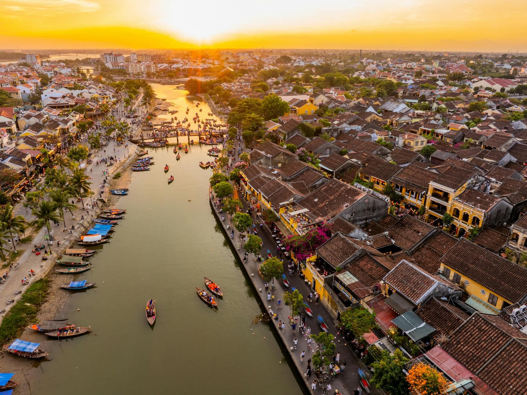 Aerial sunset view of Hoi An Ancient Town and the Thu Bon River with tourist boats lining the bank, the 2026 overtourism reality from above
