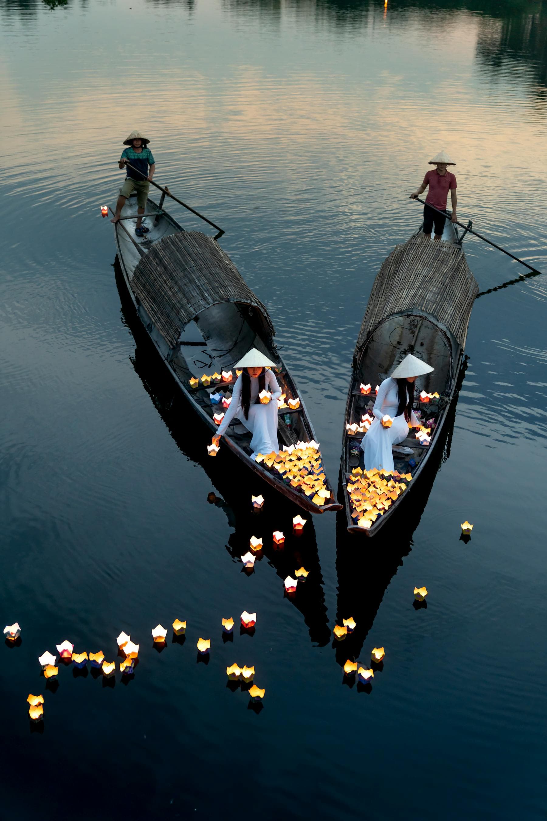 Two women in ao dai release floating candle lanterns from sampan boats onto the dark Thu Bon River at dusk in Hoi An, a 400-year-old noctourism ritual