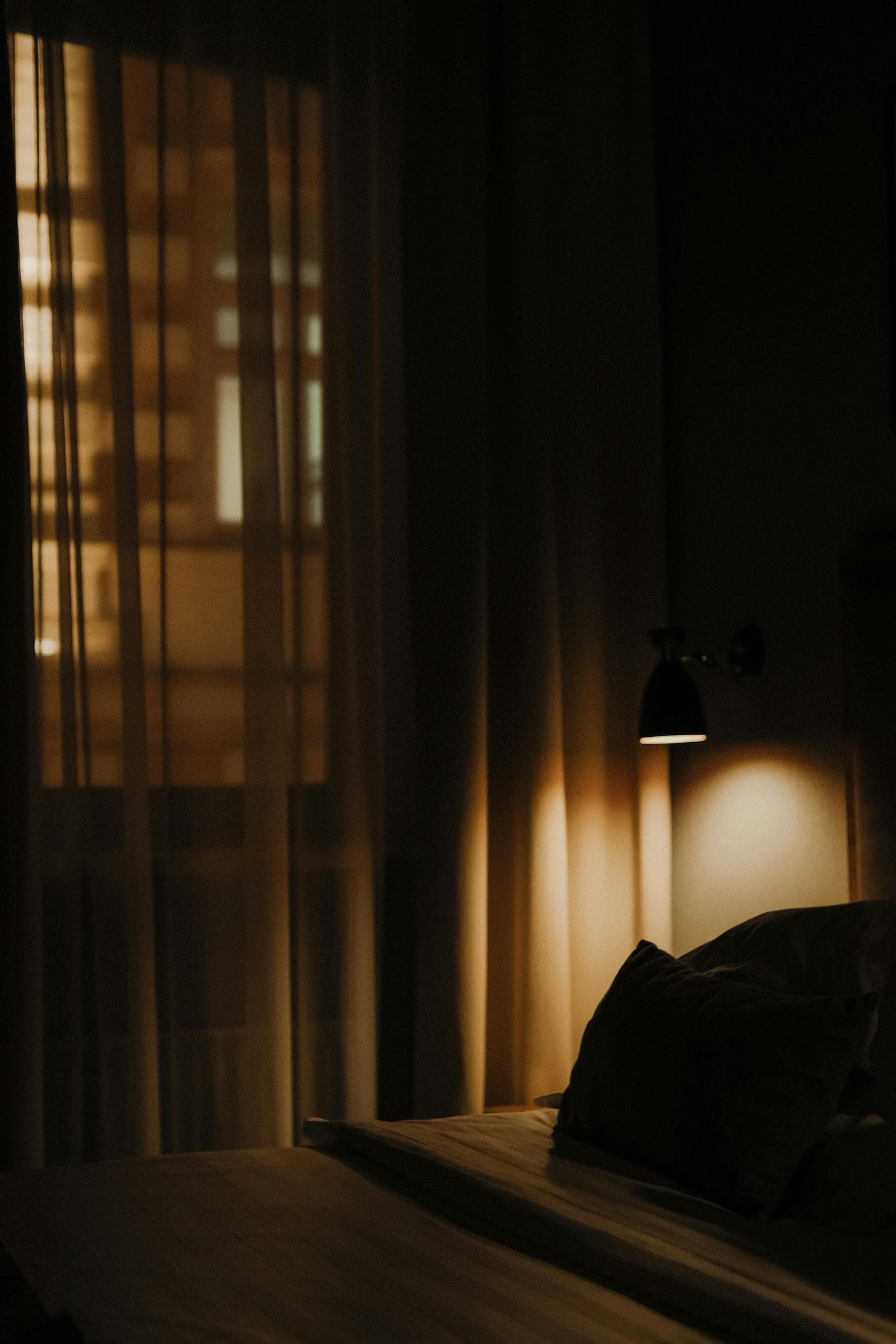 Dark hotel bedroom at night with sheer blackout curtains and a warm dim sconce, the cortisol-lowering sleep tourism environment Nghe Prana engineers for sleepmaxxing guests in Hoi An