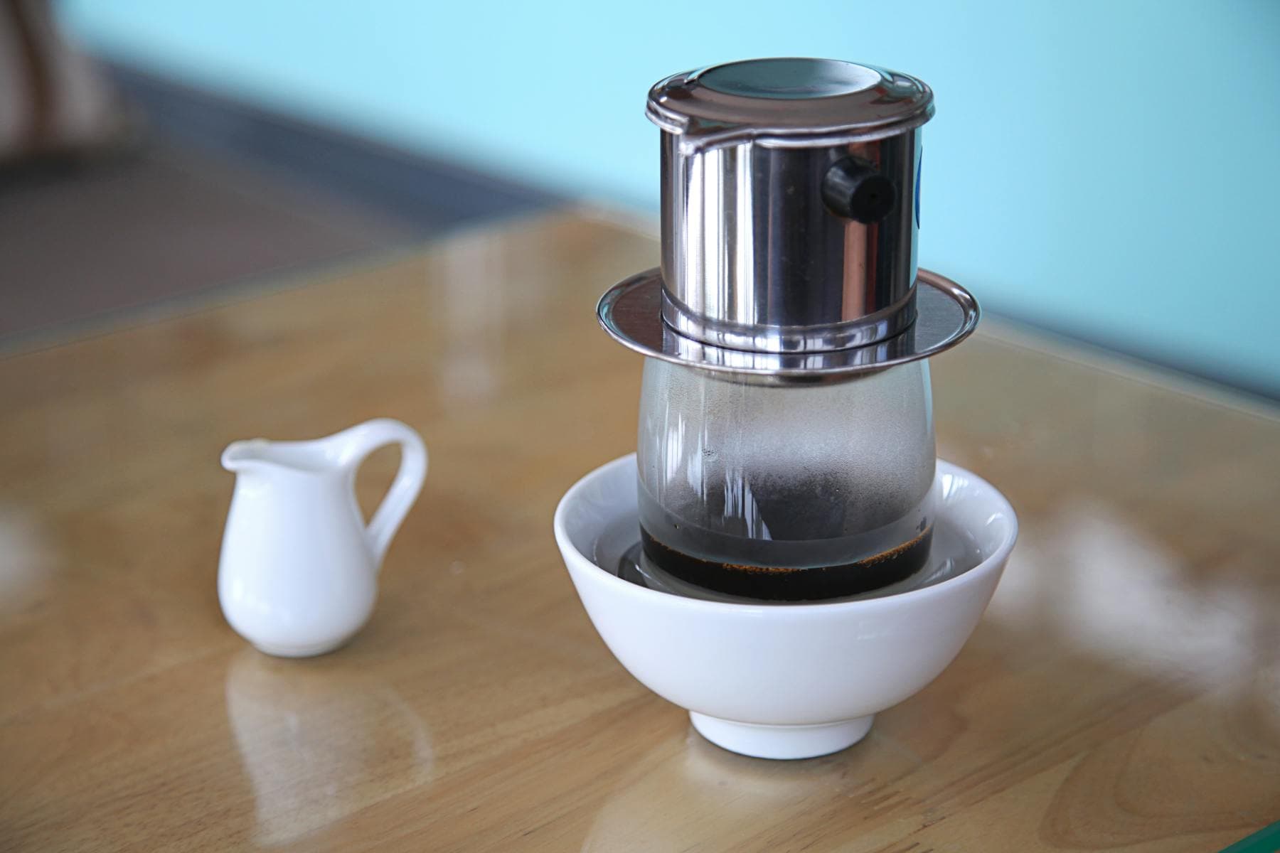 Filtre phin vietnamien traditionnel laissant tomber un café noir dans un verre, à côté d'un petit pot de lait blanc sur une table en bois — préparation classique du cà phê sữa đá